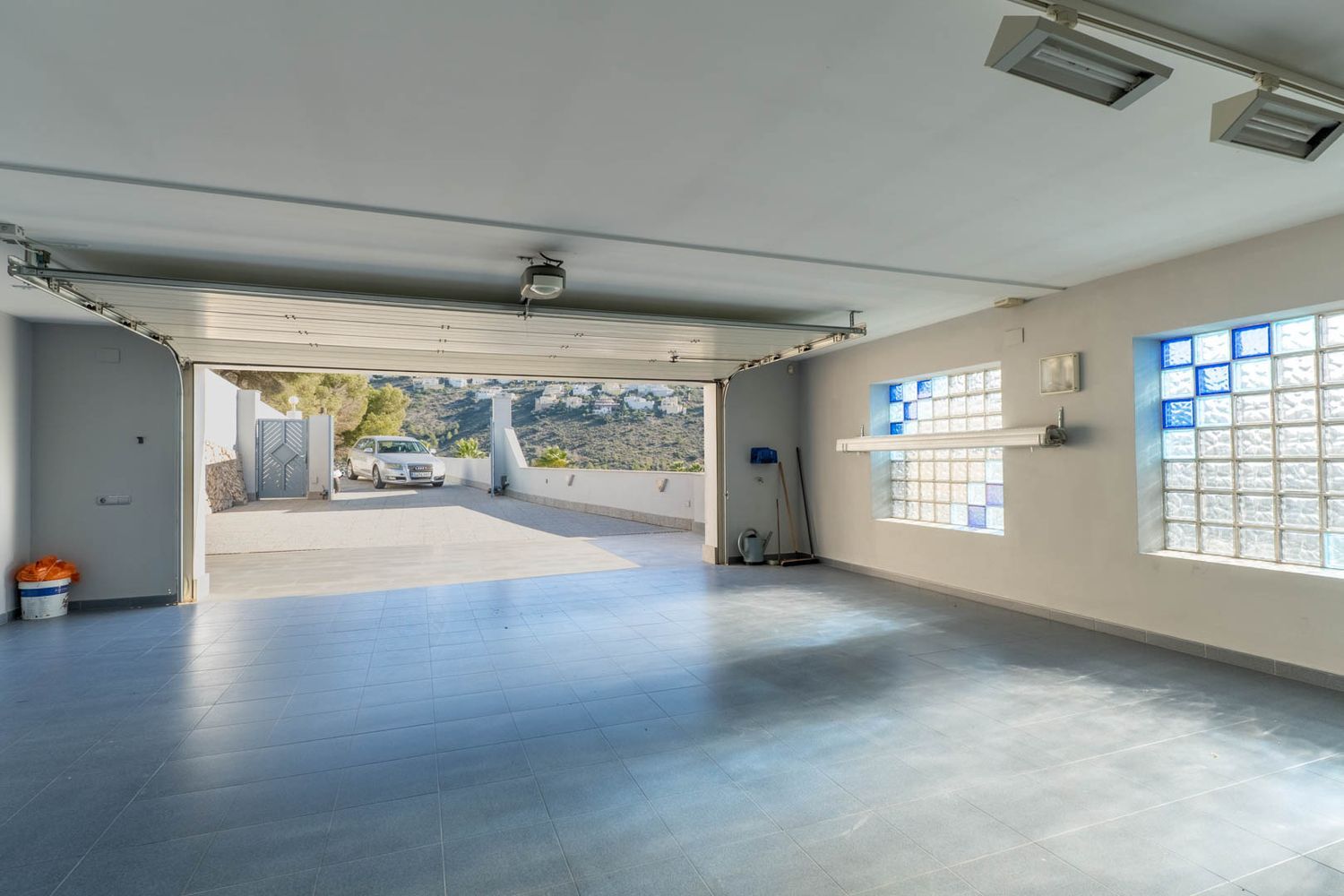 Ruime garage met lichtblauwe tegelvloer en glazen bouwstenen ramen met blauwe accenten, de deur opent naar de oprit en verre heuvels.