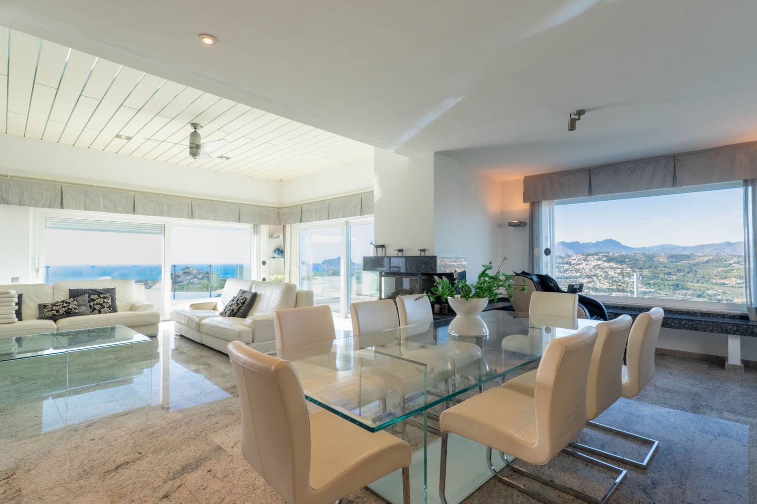 Ruime open woon- en eetkamer met gepolijste stenen vloeren. Voorzien van een glazen eettafel, witte leren banken en moderne open haard, met natuurlijk licht en uitzicht op zee en bergen.