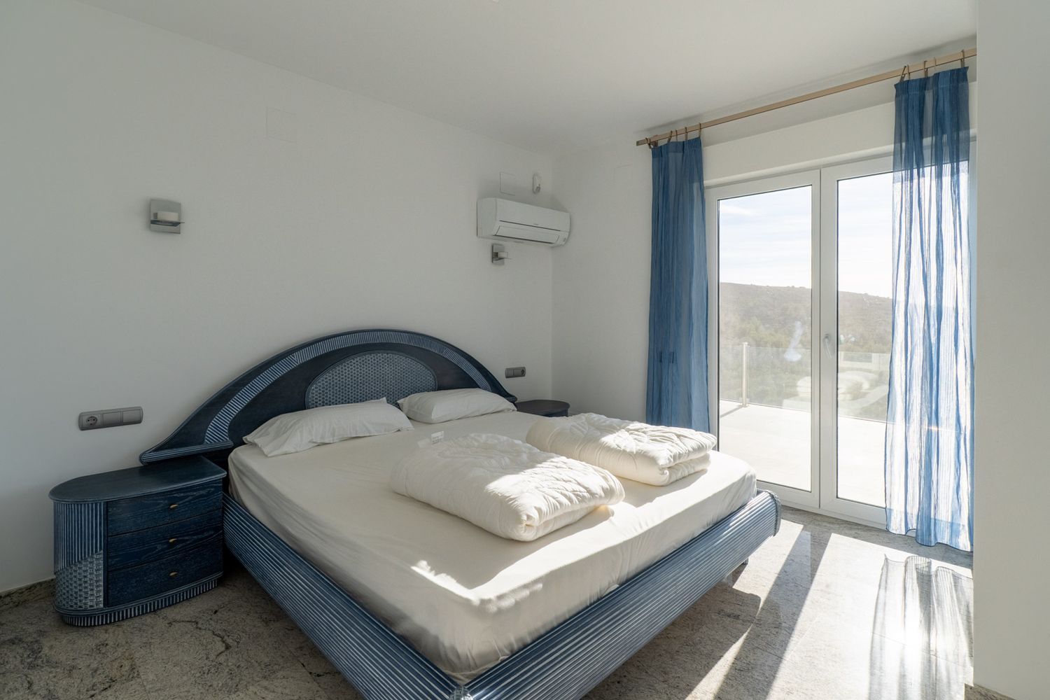 Slaapkamer met blauw designbed en bijpassende nachtkastjes, lichte vloer en directe toegang tot terras via glazen deur, met uitzicht op de heuvel en natuurlijk licht.