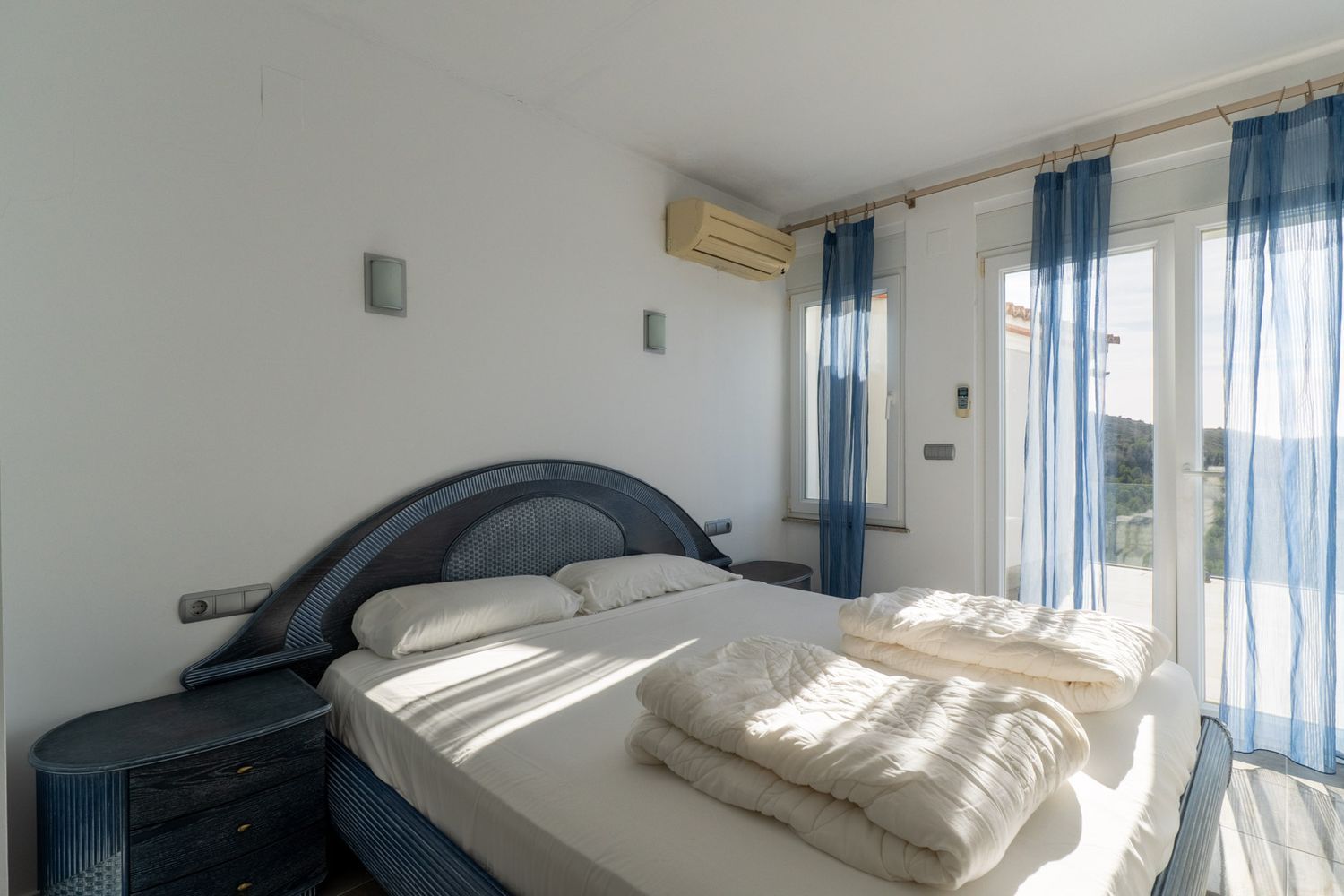 Slaapkamer met blauw-donkerhouten tweepersoonsbed en bijpassende nachtkastjes. Glazen deuren met blauwe gordijnen leiden naar een terras; airconditioning aanwezig.