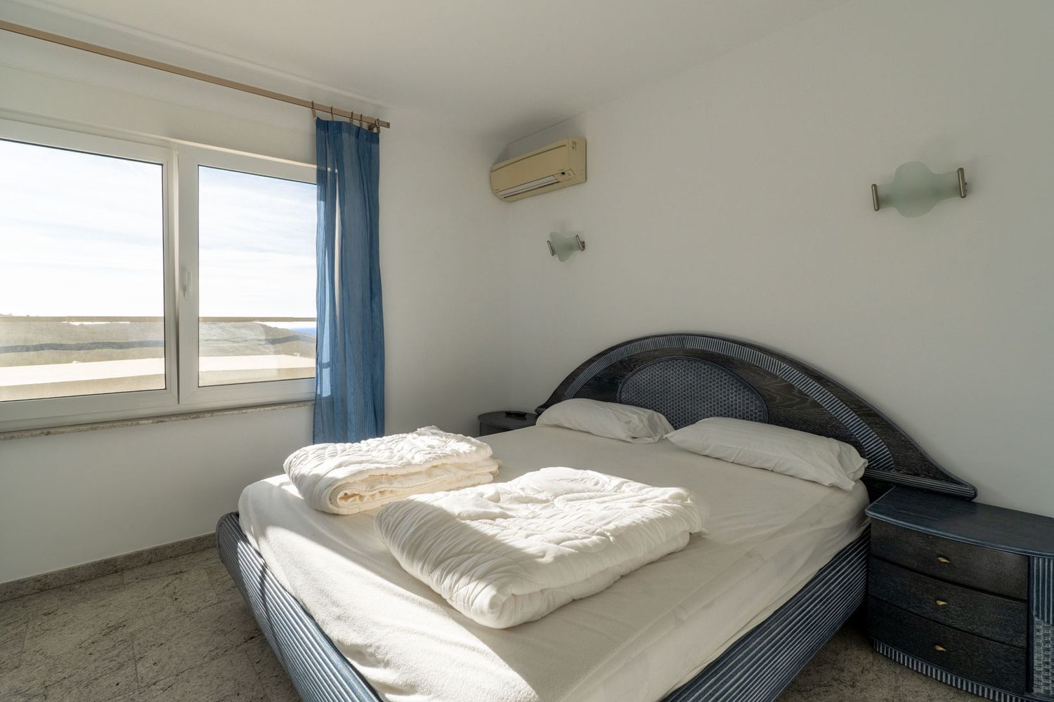 Slaapkamer met blauw gedessineerd bed en gevlochten hoofdbord, lichte tegelvloer en uitzicht naar buiten via het raam. Voorzien van airconditioning en een bijpassend nachtkastje.