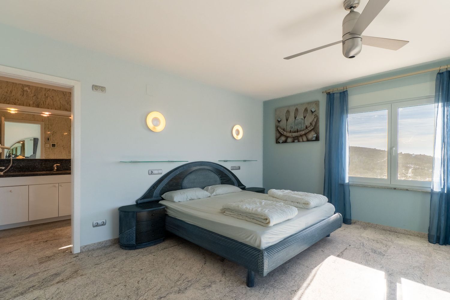 Slaapkamer met een blauwgetint designbed, bijpassende nachtkastjes en lichte marmeren vloer. Het raam biedt heuvelzicht, aangevuld met blauwe gordijnen en een zichtbare en-suite badkamer.