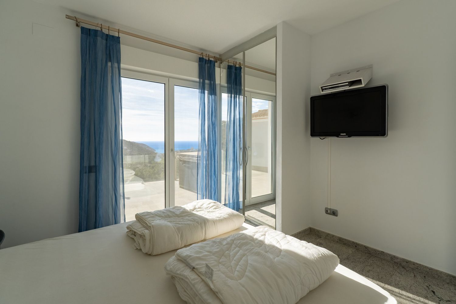 Slaapkamer met wit opgemaakt bed, wand-tv en grote glazen schuifdeuren met blauwe vitrage, die uitkomen op een terras met panoramisch uitzicht op zee.