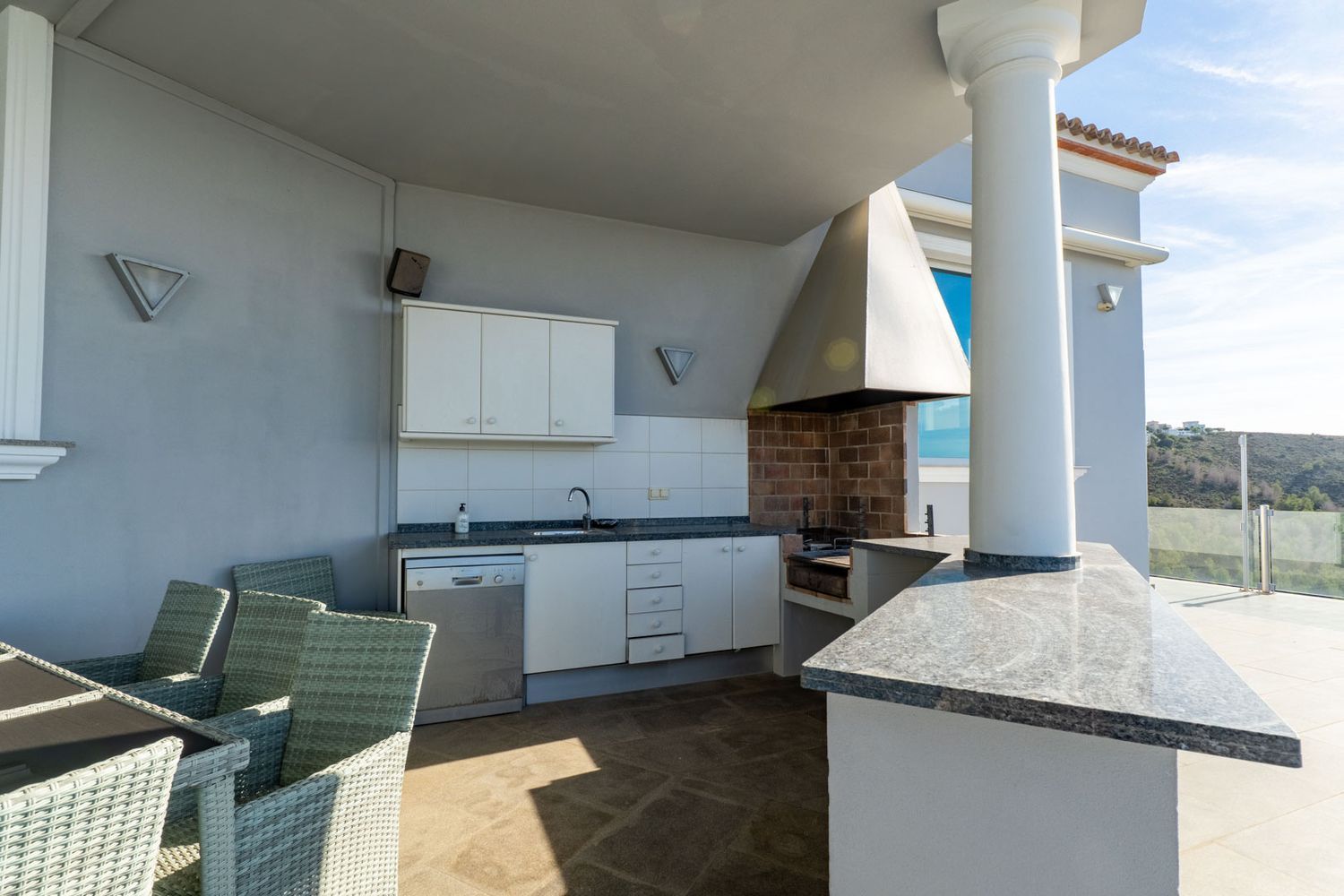 Uitgeruste buitenkeuken met bakstenen barbecue, granieten werkbladen, spoelbak en vaatwasser, grenzend aan een eettafel op een overdekt terras met uitzicht op de heuvels.
