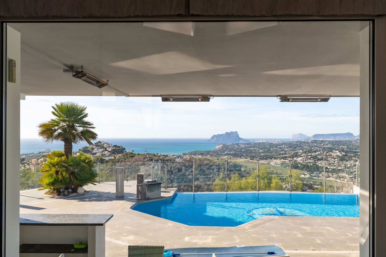 Uitzicht vanuit binnen op het terras met zwembad, palmbomen en een breed panorama van de zee, de kust van Moraira en de Peñón de Ifach. Plafondverwarming onder de overkapping.