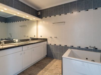 Badkamer met dubbele wastafel, donker granieten blad en witte kasten. Voorzien van een inbouwbad en een grote verlichte spiegel, met grijs-witte tegels.