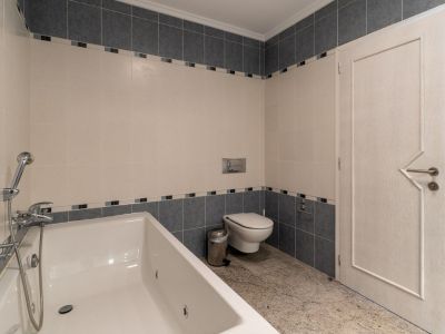 Badkamer met wit ligbad en geïntegreerde douche, wanden betegeld in beige en grijstinten met decoratieve sierrand. Lichte granieten vloer, zwevend toilet en een deur met geometrisch patroon.