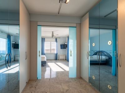 Lichte gang met granieten vloer en lichte houten kasten. Glazen schuifdeuren omlijsten een slaapkamer met zeezicht, terwijl spiegelpanelen een andere blauwgetinte slaapkamer weerspiegelen.