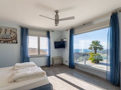 Lichte slaapkamer met groot panoramisch raam, direct zeezicht en een palmboom. Lichte graniet-effect tegelvloer en blauwe vitrage.