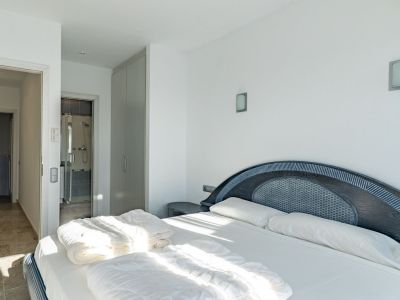 Lichte slaapkamer met tweepersoonsbed en donkerhouten hoofdbord met gevlochten inleg. Voorzien van een inbouwkast en directe toegang tot een badkamer met glazen douche.