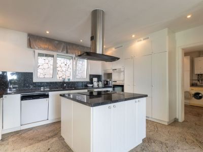Moderne keuken met witte kasten, donkere granieten werkbladen en kookeiland met geïntegreerde kookplaat. Inbouwapparatuur, uitzicht op stenen muur vanuit raam, directe toegang tot wasruimte.
