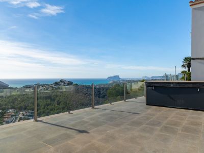 Ruim buitenterras met grijze tegels en een overdekte jacuzzi, omkaderd door een glazen balustrade met panoramisch uitzicht op zee en de Peñón de Ifach.