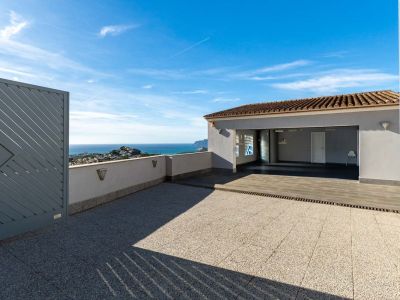 Ruime buitenruimte met tegelbestrating en toegang tot een overdekte garage. Biedt panoramisch uitzicht op de Middellandse Zee en de kust, omkaderd door een modern grijs hek en een lage muur met verlichting.