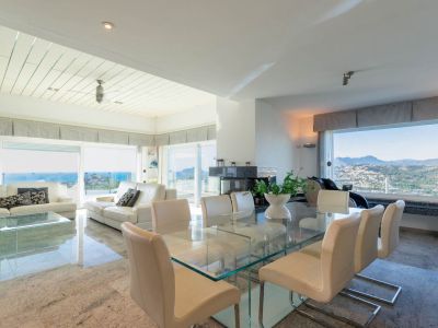 Ruime open woon- en eetkamer met gepolijste stenen vloeren. Voorzien van een glazen eettafel, witte leren banken en moderne open haard, met natuurlijk licht en uitzicht op zee en bergen.