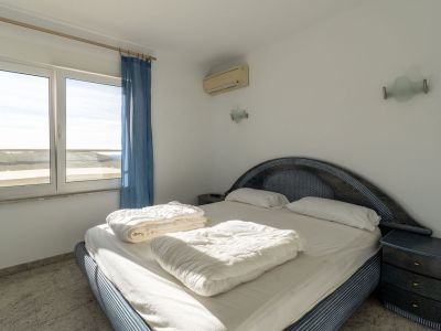 Slaapkamer met blauw gedessineerd bed en gevlochten hoofdbord, lichte tegelvloer en uitzicht naar buiten via het raam. Voorzien van airconditioning en een bijpassend nachtkastje.
