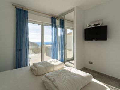 Slaapkamer met wit opgemaakt bed, wand-tv en grote glazen schuifdeuren met blauwe vitrage, die uitkomen op een terras met panoramisch uitzicht op zee.