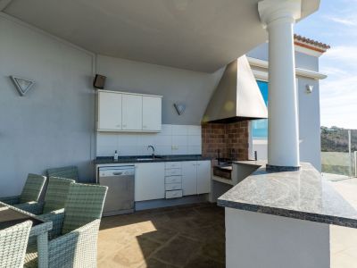 Uitgeruste buitenkeuken met bakstenen barbecue, granieten werkbladen, spoelbak en vaatwasser, grenzend aan een eettafel op een overdekt terras met uitzicht op de heuvels.