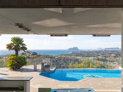 Uitzicht vanuit binnen op het terras met zwembad, palmbomen en een breed panorama van de zee, de kust van Moraira en de Peñón de Ifach. Plafondverwarming onder de overkapping.