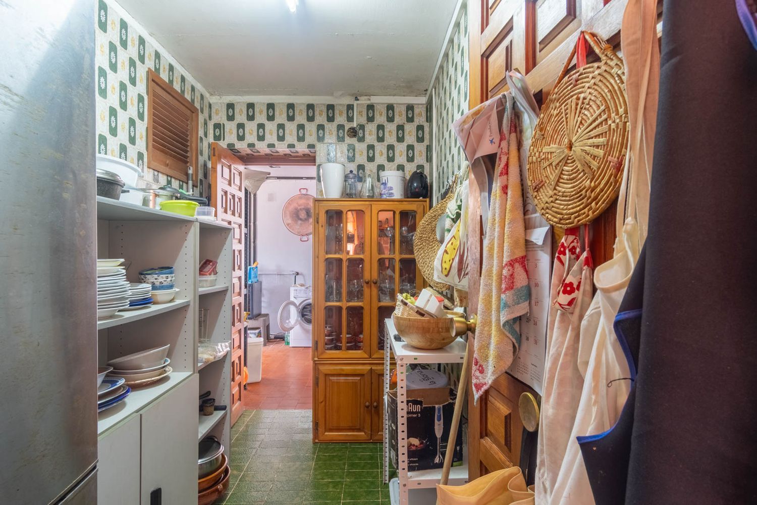 Functionele bijkeuken met patroonbehang, groene tegelvloer, open schappen met keukengerei en een houten vitrinekast. Een wasruimte is achterin zichtbaar.