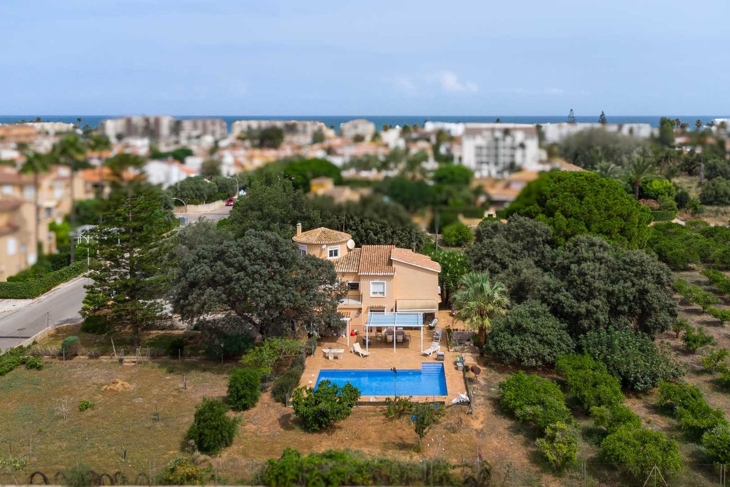 Luchtfoto van de villa met blauw zwembad, een schaduwrijk terras en een ruime, groene tuin. Op de achtergrond zijn de stad en de Middellandse Zee zichtbaar.