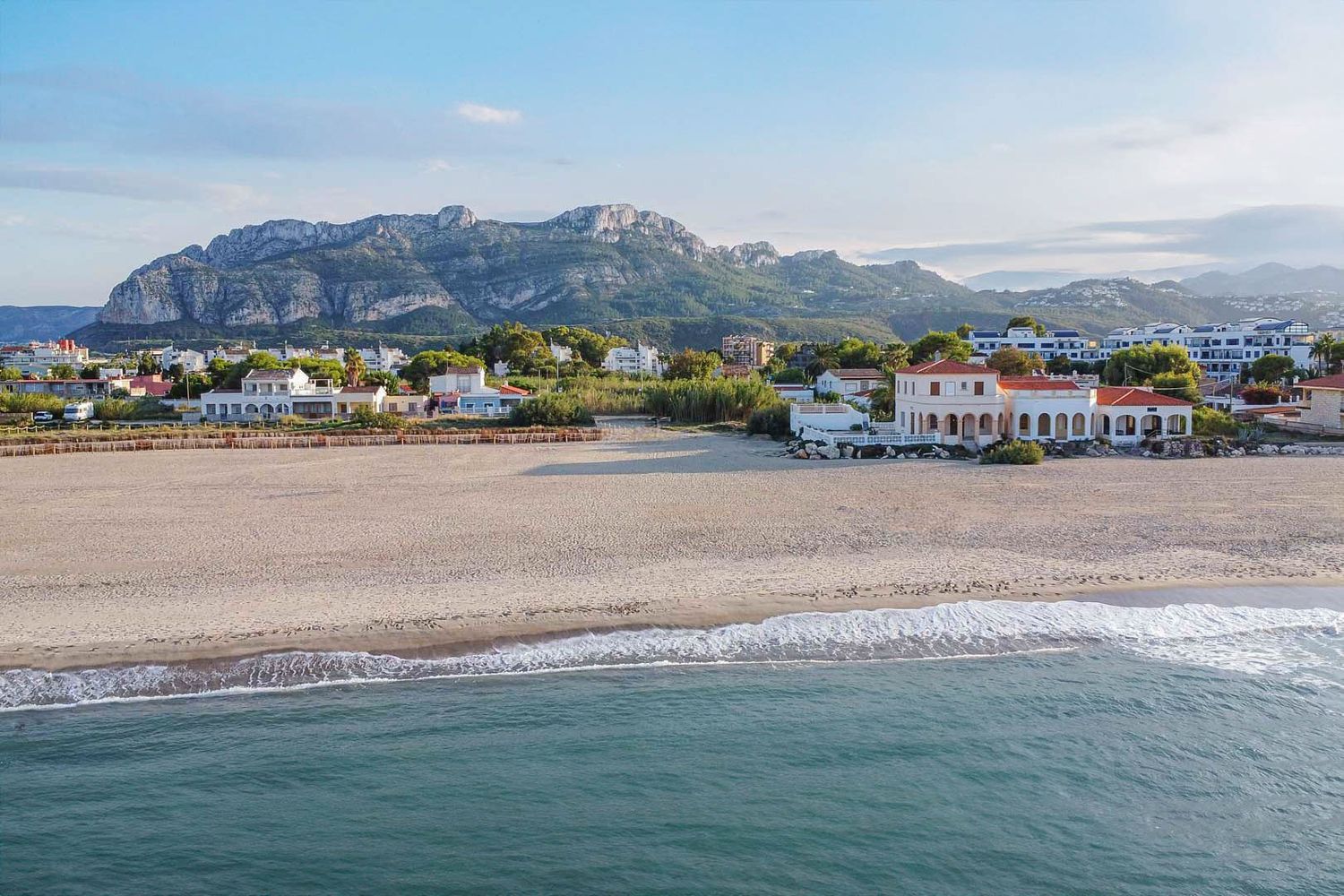 Luchtfoto van het zandstrand met zachte golven, met een klassiek kusthuis met bogen, tegen de achtergrond van het Montgó-massief.