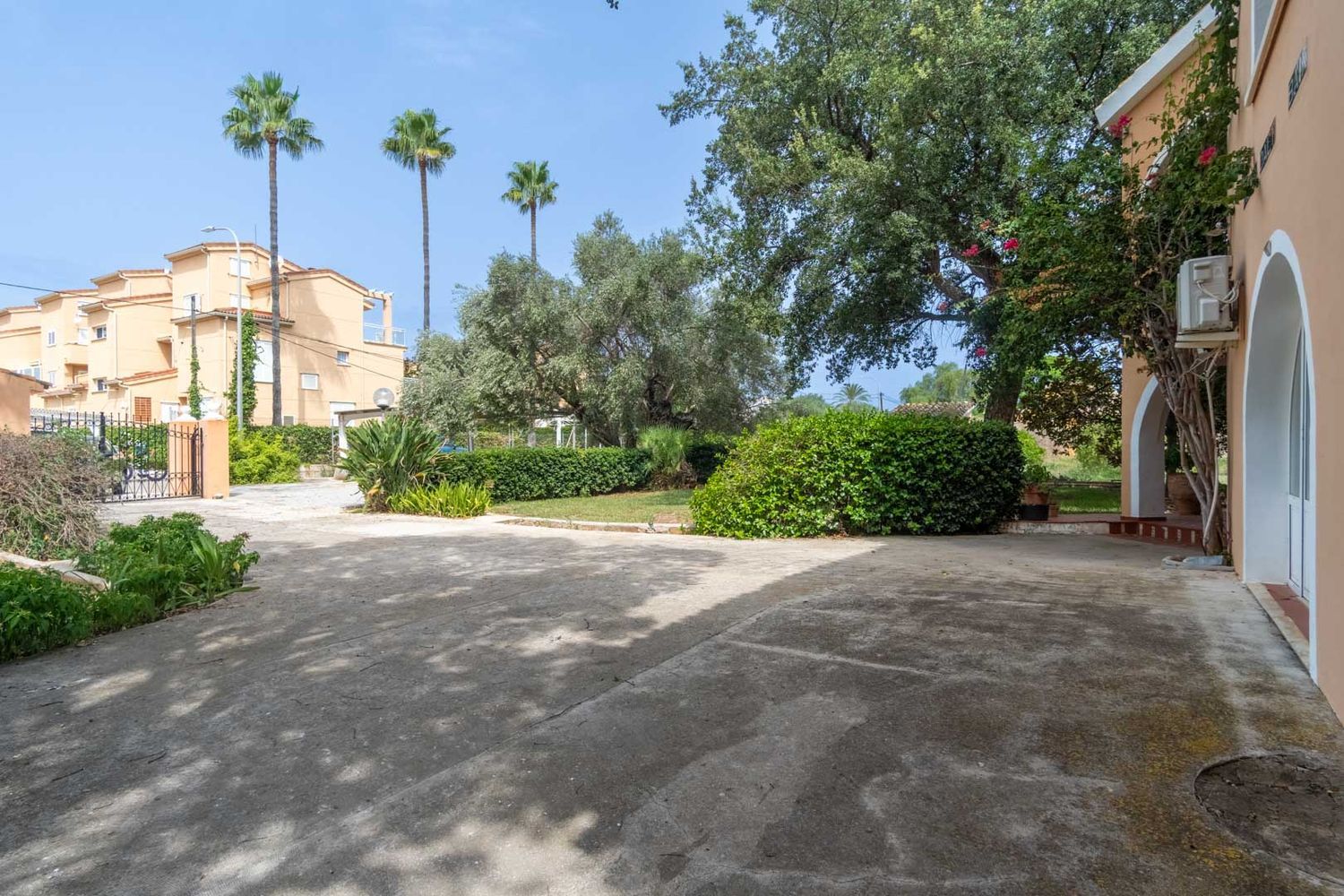 Ruime betonnen privéparkeerplaats, begrensd door een mediterrane tuin met palmbomen, olijfbomen en struiken. Een deel van de gebogen gevel van het chalet is zichtbaar.