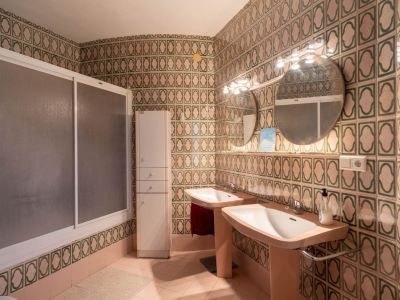 Badkamer met opvallende groen-beige wandtegels, dubbele wastafel met twee wasbakken, ronde spiegels met verlichting, douchecabine en een hoge opbergkast.