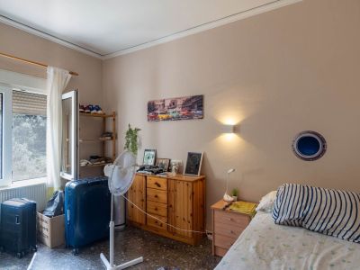 Functionele slaapkamer met eenpersoonsbed, houten nachtkastje en ladekast, verlicht door natuurlijk licht van het raam en een wandlamp. Donkere, gespikkelde vloer.