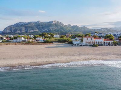 Luchtfoto van het zandstrand met zachte golven, met een klassiek kusthuis met bogen, tegen de achtergrond van het Montgó-massief.