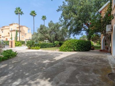 Ruime betonnen privéparkeerplaats, begrensd door een mediterrane tuin met palmbomen, olijfbomen en struiken. Een deel van de gebogen gevel van het chalet is zichtbaar.