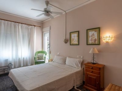 Slaapkamer met tweepersoonsbed, houten nachtkastje en lamp. Voorzien van een plafondventilator, groene rieten stoel en glazen deur naar buiten.