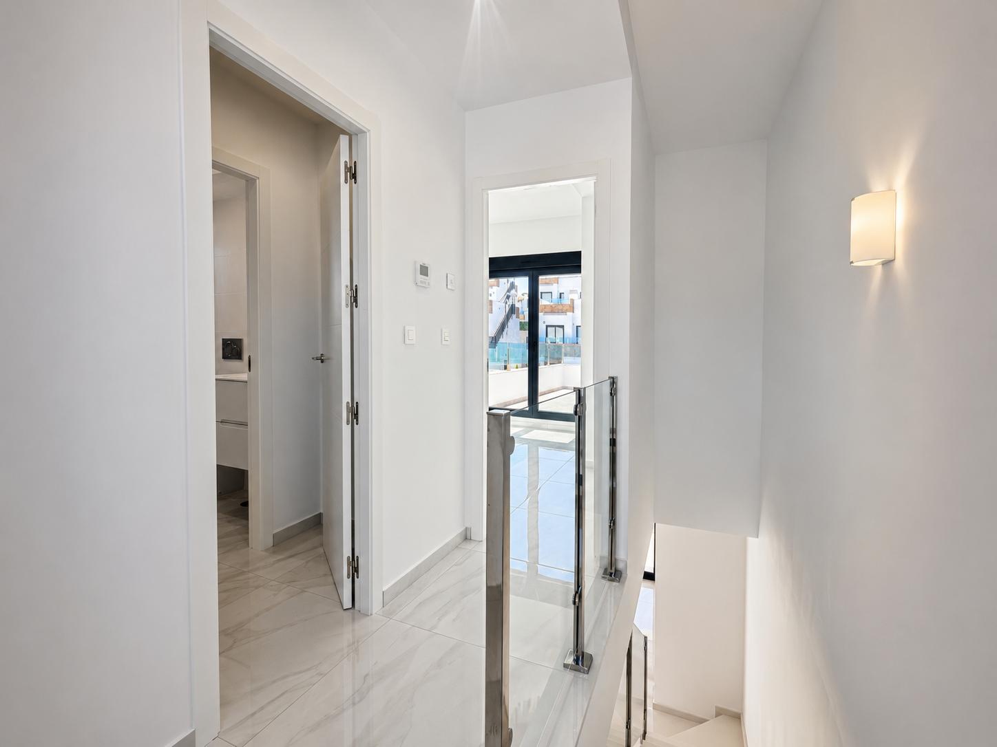 Lichte bovenverdieping hal met gepolijste marmerlook vloertegels en moderne glazen en stalen trapleuning. Een deur leidt naar een badkamer, een andere naar een buitenbalkon met daglicht.