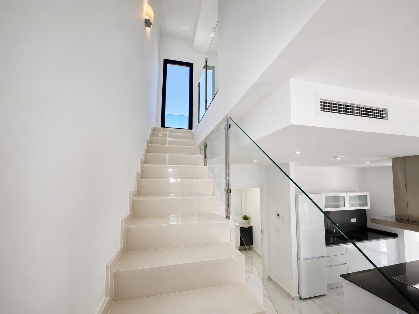 Moderne trap met lichte treden en een strakke glazen balustrade, leidend naar een buitendeur. Een eigentijdse keuken met zwarte werkbladen en een glimp van een badkamer zijn zichtbaar.