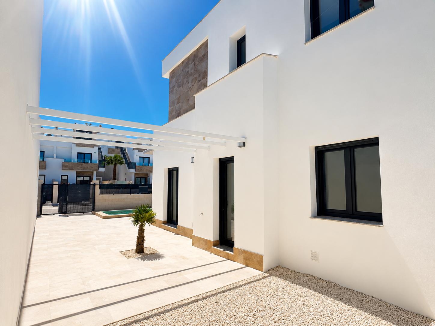Moderne villa-exterieur met witte gevel en steenaccenten. Lichte tegelterras, kleine palmboom, witte pergola en donker omkaderde ramen onder felle zon.