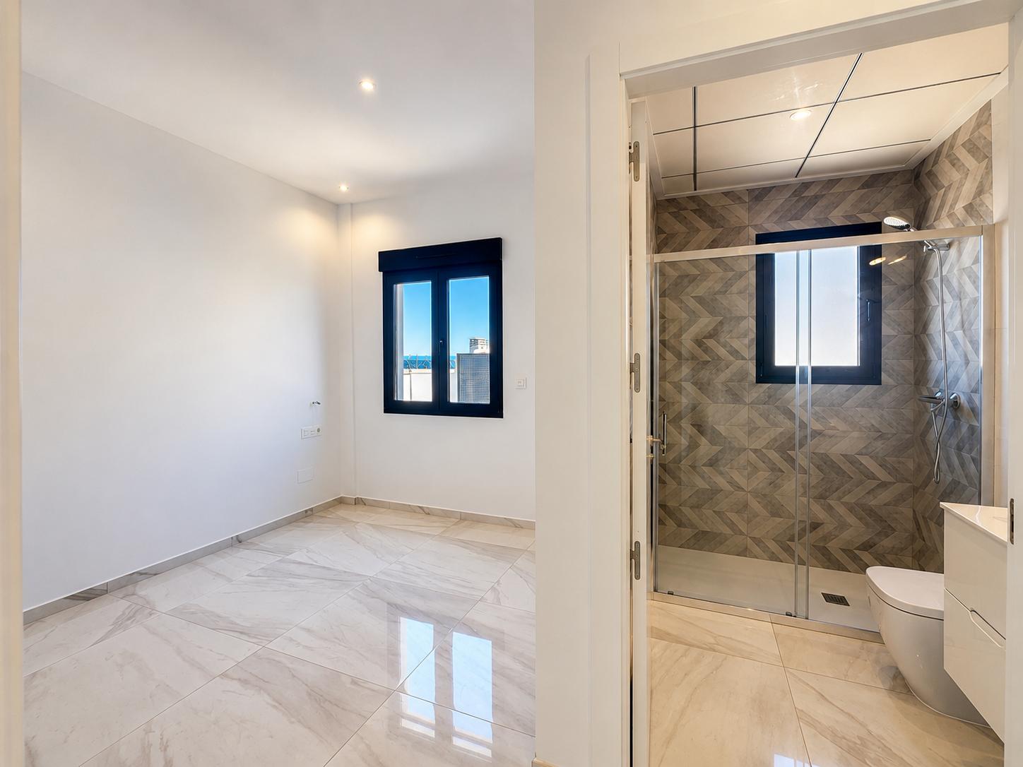 Ruime slaapkamer met lichte marmerlook vloer en donker kozijnraam met verre uitzichten, grenzend aan een en-suite badkamer met geometrische douchewandtegels.