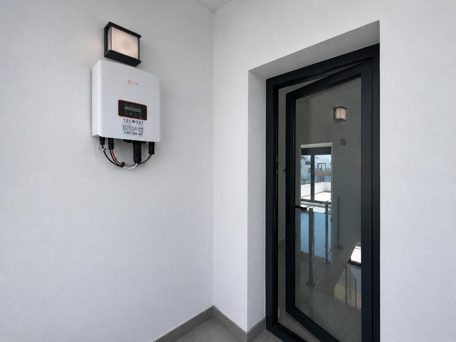 Solis omvormer voor zonne-energie op witte muur met buitenlamp. Een deels geopende zwartomlijste glazen deur toont een binnentrap met balustrade.