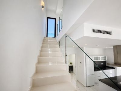 Moderne trap met lichte treden en een strakke glazen balustrade, leidend naar een buitendeur. Een eigentijdse keuken met zwarte werkbladen en een glimp van een badkamer zijn zichtbaar.