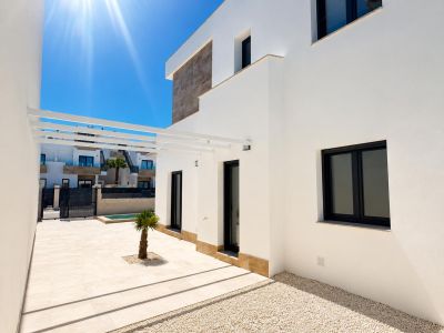 Moderne villa-exterieur met witte gevel en steenaccenten. Lichte tegelterras, kleine palmboom, witte pergola en donker omkaderde ramen onder felle zon.