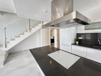 Open keuken met donker aanrechtblad eiland en witte inductiekookplaat, grenzend aan een moderne trap met glazen balustrade en witte treden.