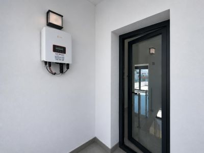 Solis omvormer voor zonne-energie op witte muur met buitenlamp. Een deels geopende zwartomlijste glazen deur toont een binnentrap met balustrade.