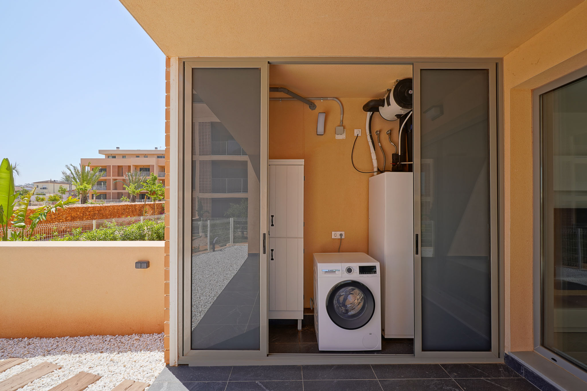 Buitenwasruimte met Bosch wasmachine en aerothermisch systeem, bereikbaar via een betegeld terras met siergrind en tuinzicht.