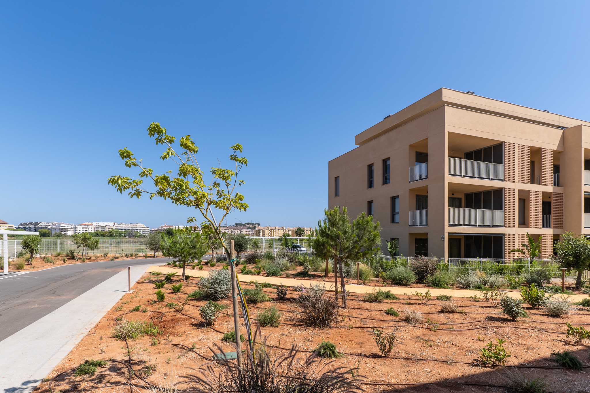Exterieur van het moderne woongebouw met zandkleurige gevel en balkons met witte balustrades. De jonge mediterrane aanleg grenst aan een geasfalteerde toegangsweg.