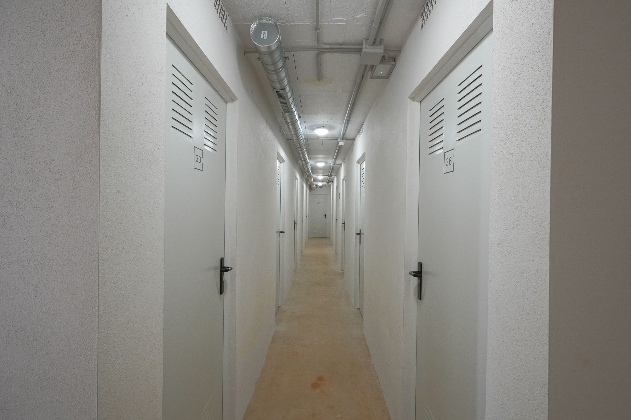 Functionele gang naar opslagruimtes, met genummerde witte deuren en ventilatiesleuven. Zichtbetonplafond met industriële leidingen en inbouwverlichting, aangevuld met gestructureerde muren.