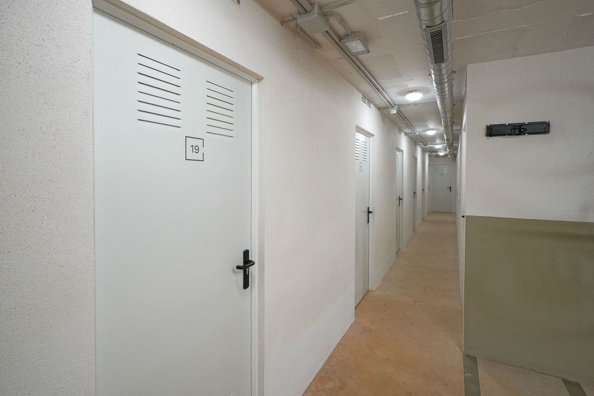 Gang met witte opslagdeuren voorzien van ventilatieroosters en zwarte handgrepen. Het plafond toont zichtbare leidingen en inbouwverlichting boven een betonnen vloer.