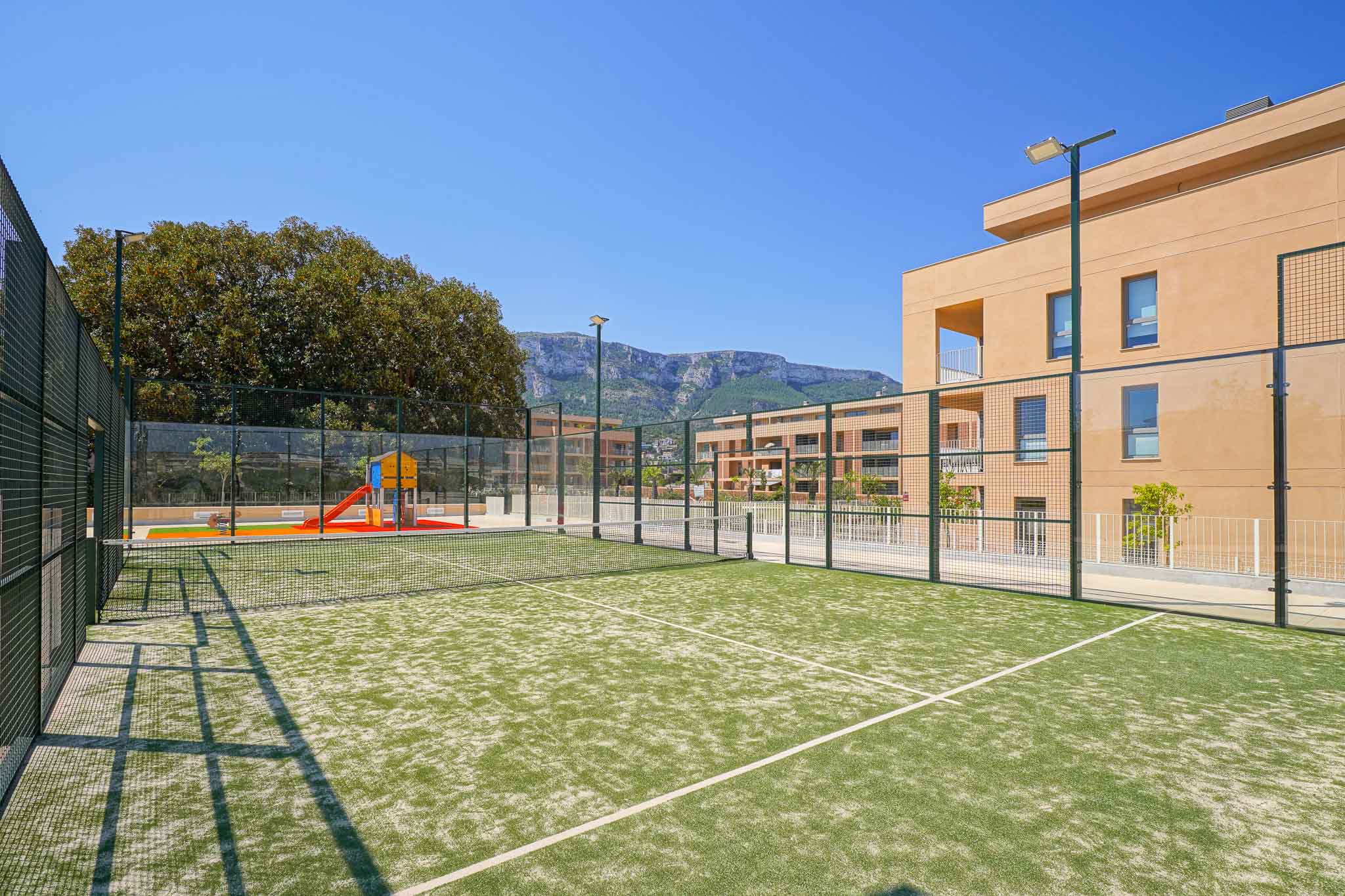 Groene kunstgras padelbaan, omheind en voorzien van verlichting, grenzend aan een kinderspeelplaats en moderne woongebouwen met uitzicht op de bergen.