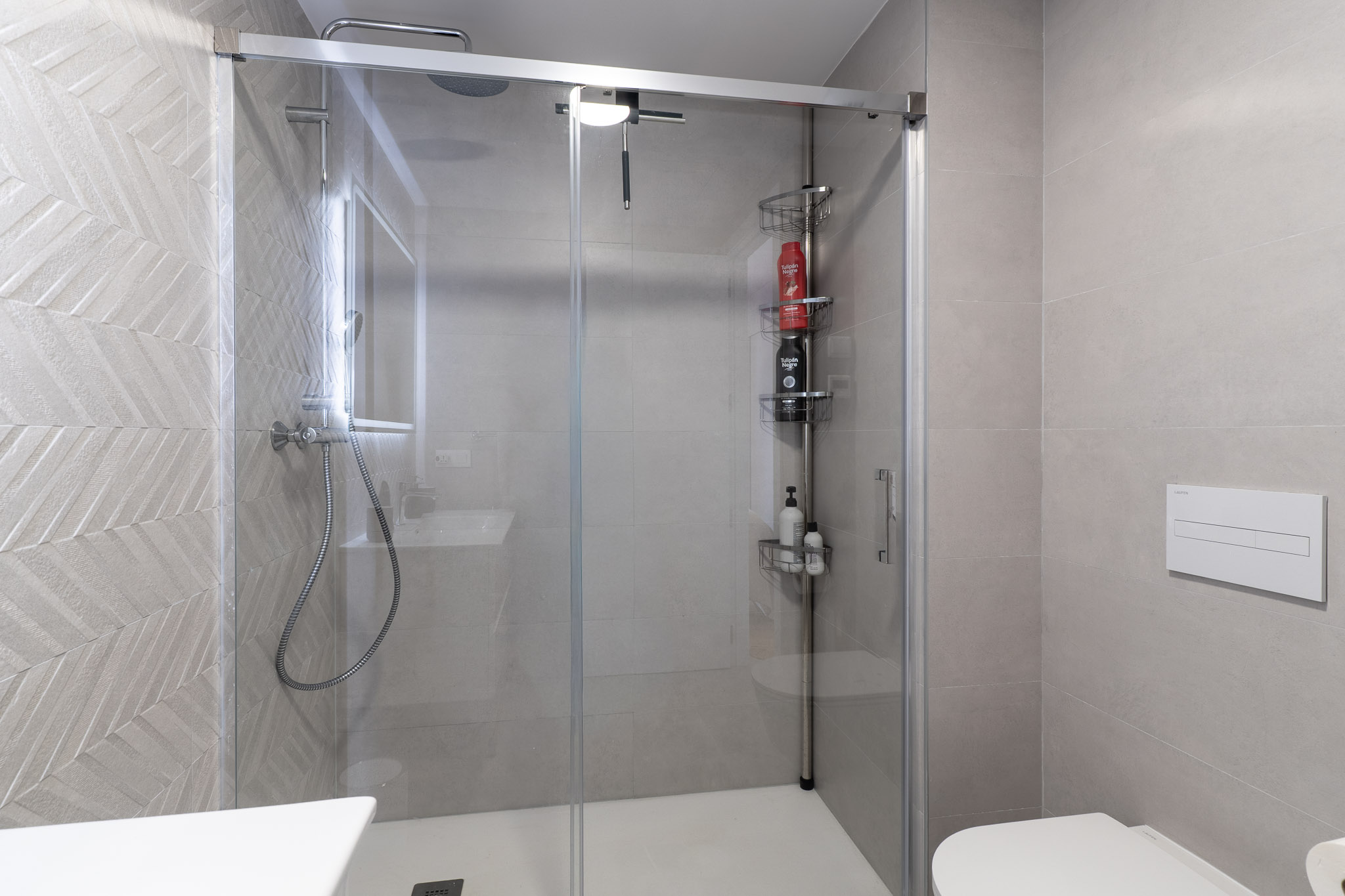 Hedendaagse badkamer met glazen douchecabine en witte douchebak. De douchewand is voorzien van lichtgrijze tegels met een visgraatpatroon, aangevuld met een regendouche en zwevend toilet.