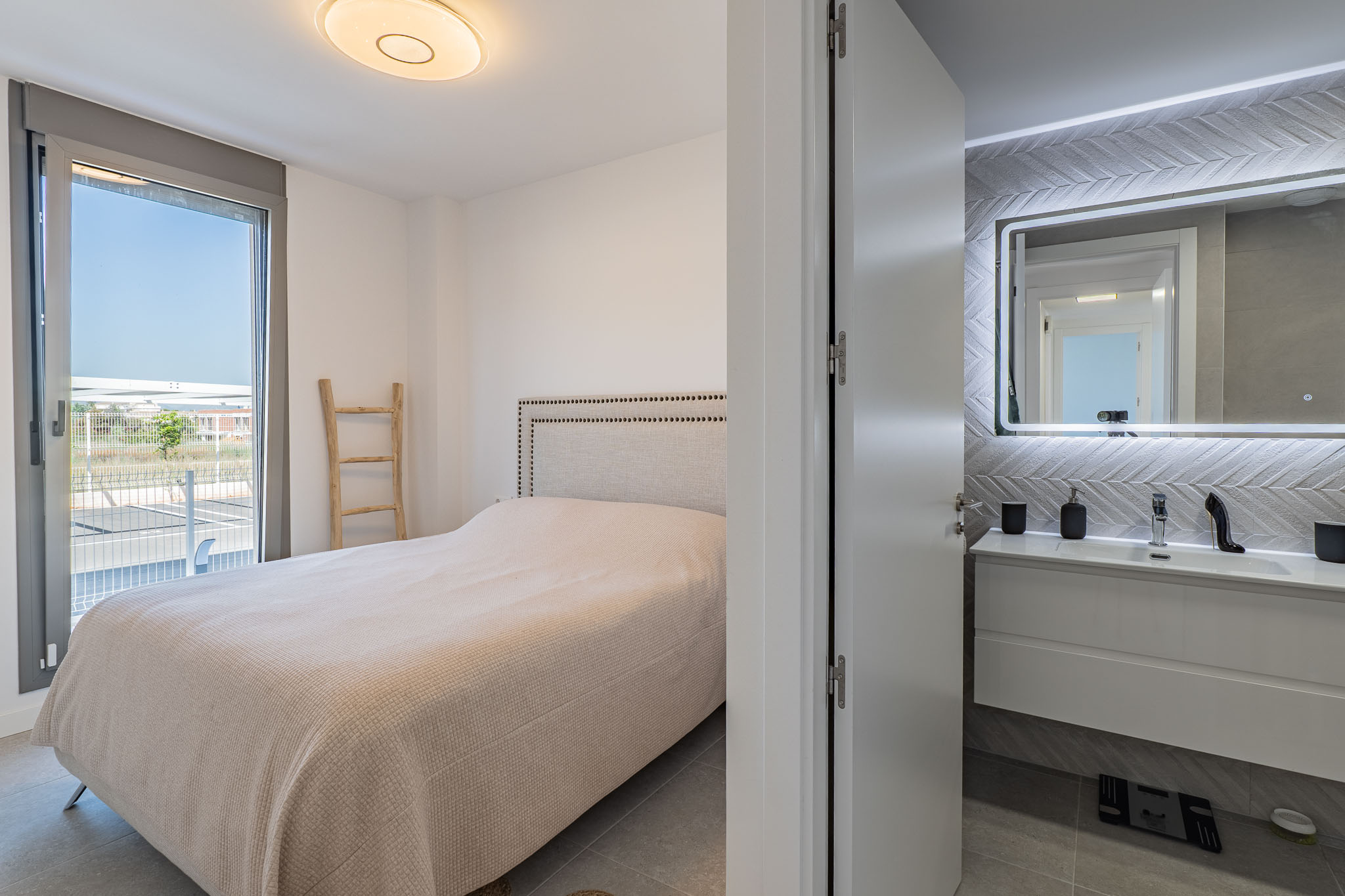 Hoofdslaapkamer met gestoffeerd hoofdbordbed en directe toegang tot een buitenterras. Een en-suite badkamer met zwevend wastafelmeubel en verlichte spiegel is zichtbaar.