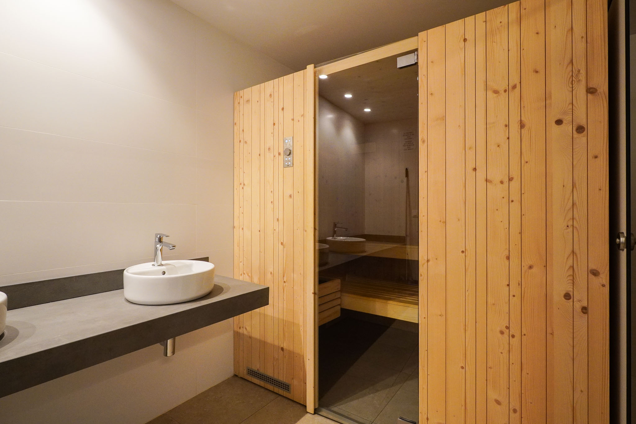 Kleedruimte met een strakke zwevende wastafel en opzetkom, naast de uitnodigende houten lamellen sauna-ingang met glazen paneel.