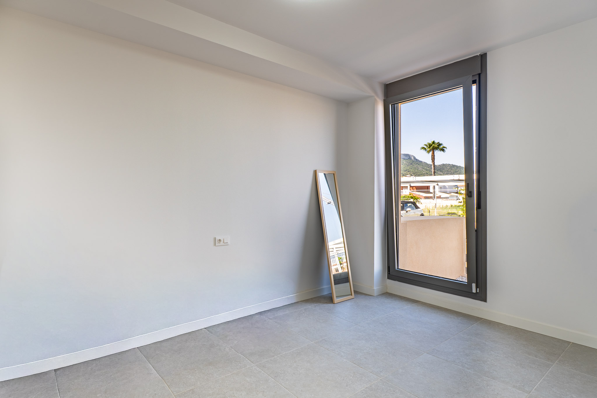 Lege kamer met witte muren en grijze tegelvloer, klaar voor inrichting. Groot raam biedt natuurlijk licht en uitzicht op een palmboom en verre bergen.