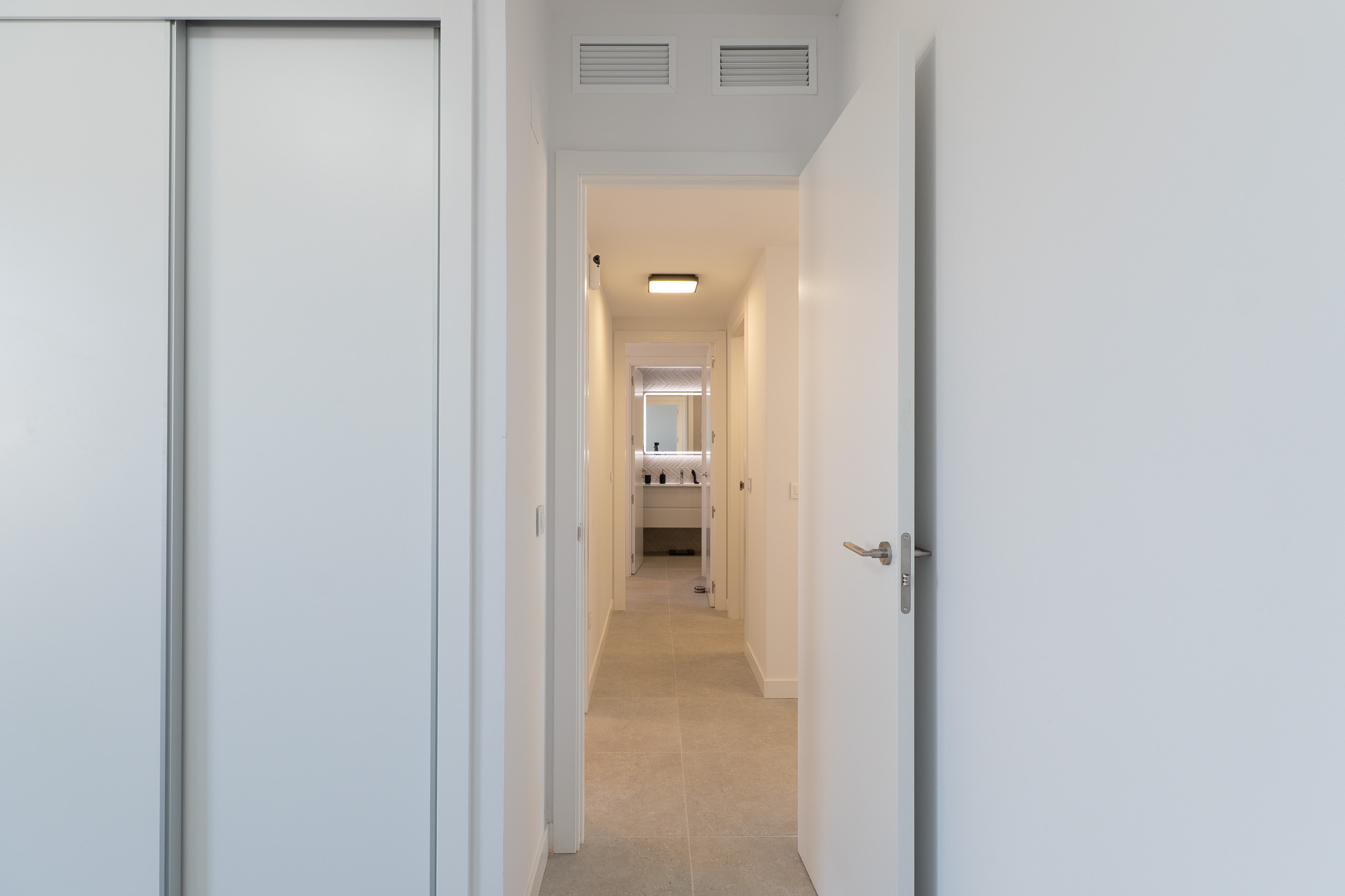 Lichte gang met witte schuifkast en open deur. Lichte vloertegels leiden naar een moderne badkamer met verlichte spiegel en eigentijds badmeubel. Ventilatieopeningen in het plafond zichtbaar.